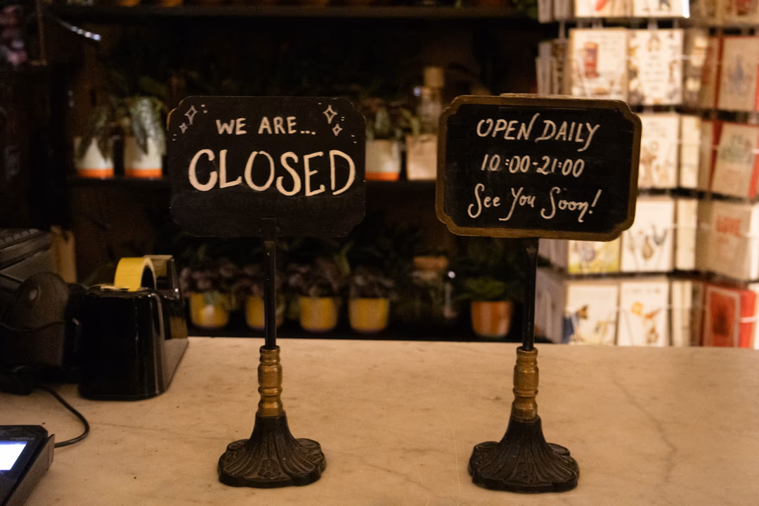 店家櫃台上並排立著兩塊手寫牌子，一塊寫著 CLOSED、一塊寫著 OPEN DAILY，象徵每個商家都要自己決定要不要對 AI 代理人開門